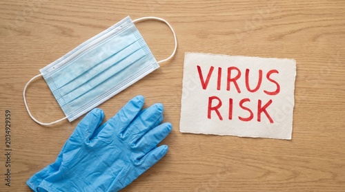 Medical face mask with blue nitrile glove and a paper sign with text virus risk on a wooden table.