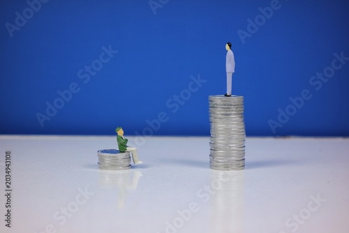 Two miniature figures stand on different stacks of coins against a dark blue background, symbolizing wealth disparity, income gap, economic inequality, finance, and social contrast.