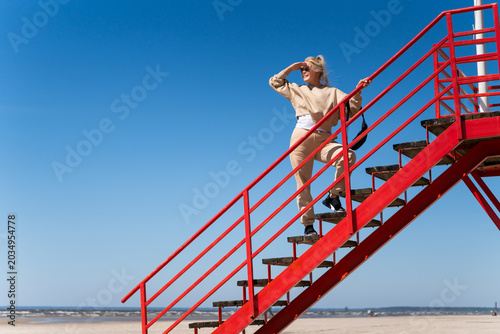 Smiling young woman in cozy casual outfit standing on red lifeguard tower stairs at Parnu beach in Estonia, enjoying sunny spring day before summer season