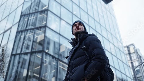 Tracking shot of young man with backpack turning and looking up beside glass office building in downtown on overcast winter morning, handheld tilt upward