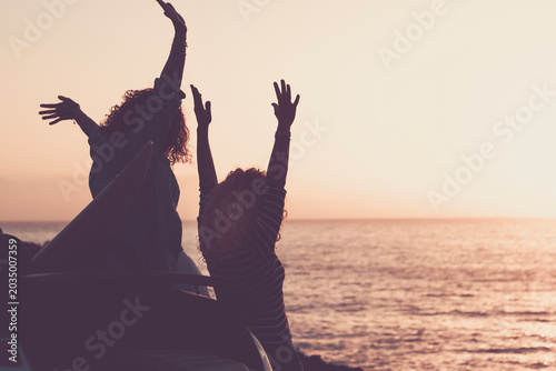 Two friends raise their arms in celebration on the roof of a car at sunset by the ocean, capturing carefree summer joy, freedom and friendship during a road trip along the coast.