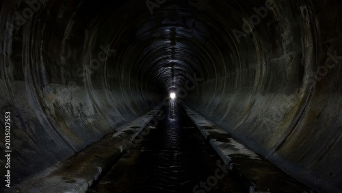  Dark concrete drainage tunnel with distant light at end. 
