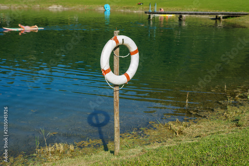 Canvas Print Lifebuoy mounted on wooden post near calm lake with swimmers and pier in backgro