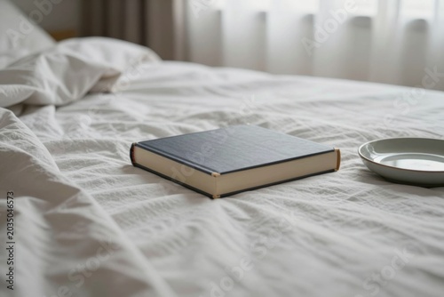 Closed black hardcover book and small ceramic plate on a white rumpled sheets. in a bedroom.