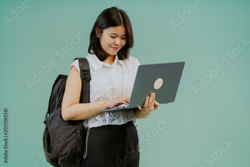 Young smiling smart asian teen girl student wears a white shirt and backpack holding use a laptop computer isolated on light blue studio background. High school, university and college concept.