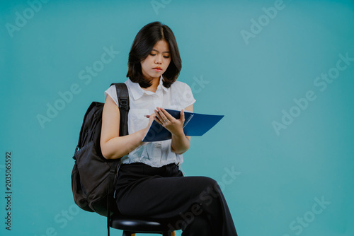 Asian female student woman taking exam wearing a white shirt and school bag sitting on the chair writing on the notebook, isolated on tosca blue background. Education learning concept.