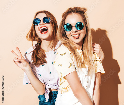 Two young beautiful smiling blond hipster in trendy summer colorful T-shirt clothes. Sexy carefree women posing near beige wall in round sunglasses. Showing peace sign