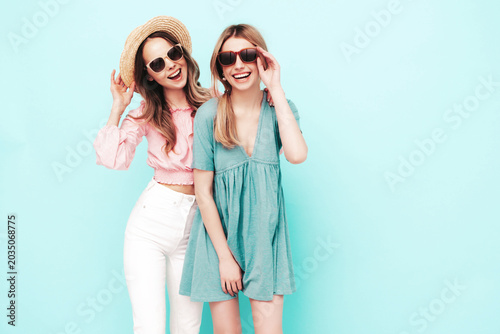 Two young beautiful smiling brunette hipster female in trendy summer dresses. Sexy carefree women posing near blue wall. Positive models having fun. Cheerful and happy. In hats and sunglasses