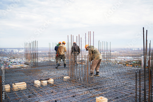 Canvas Print A team of construction workers in hard hats walk across a complex steel rebar grid with vertical dowels on a high-rise building slab overlooking a wide cityscape