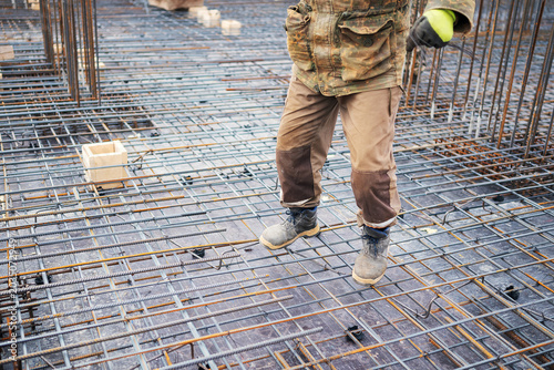 A close-up view of an ironworker's legs in reinforced trousers and safety work boots standing on a steel rebar reinforcement grid holding tying wire.