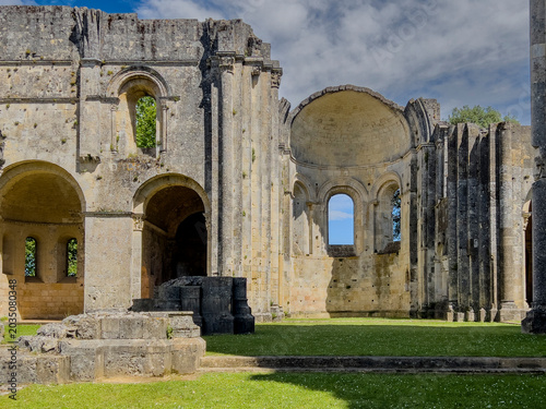 Visitors walk around the historic Abbaye de la Sauve-Majeur in Gironde, France. The ruins stand under a blue sky with clouds, revealing details of the architecture and scenery.