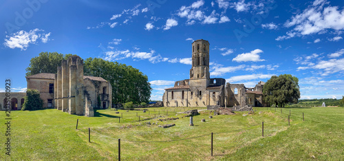 Visitors walk around the historic Abbaye de la Sauve-Majeur in Gironde, France. The ruins stand under a blue sky with clouds, revealing details of the architecture and scenery.