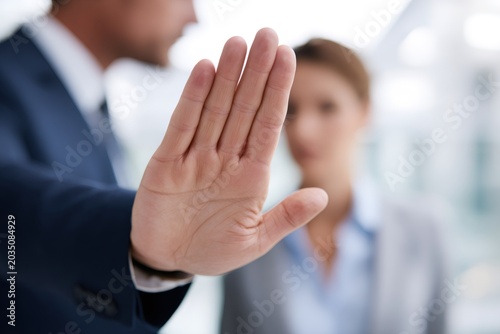 Businessman showing a stop hand gesture with a female colleague in the background. Concept of workplace conflict, rejection, or harassment prevention. Focus on hand