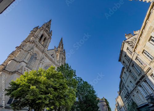 Saint-Louis Church rises above the trees in Quartier des Chartrons, Bordeaux. The church features tall spires and intricate details on its stone facade. The clear sky adds to its presence.