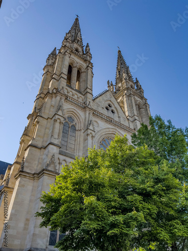 Saint-Louis Church rises above the trees in Quartier des Chartrons, Bordeaux. The church features tall spires and intricate details on its stone facade. The clear sky adds to its presence.