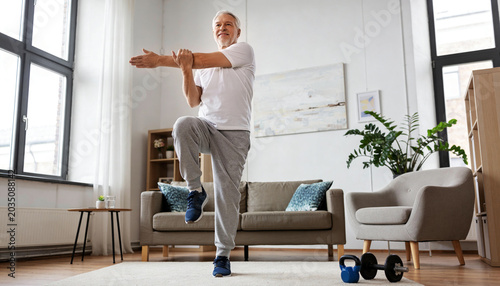 Doing balance stretch, senior man wearing exercise clothes raising arms in living room, kettlebell