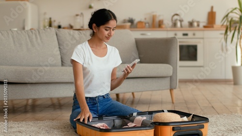 Cheerful Indian Arabian woman at home preparing trip vacation travel female girl checking items packing clothes using mobile phone online list remembering put summer hat inside luggage journey indoors