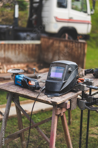 Assorted workshop tools including welding helmet and cutting discs placed on a wooden stool outdoors. Industrial DIY and metalworking equipment in natural light.