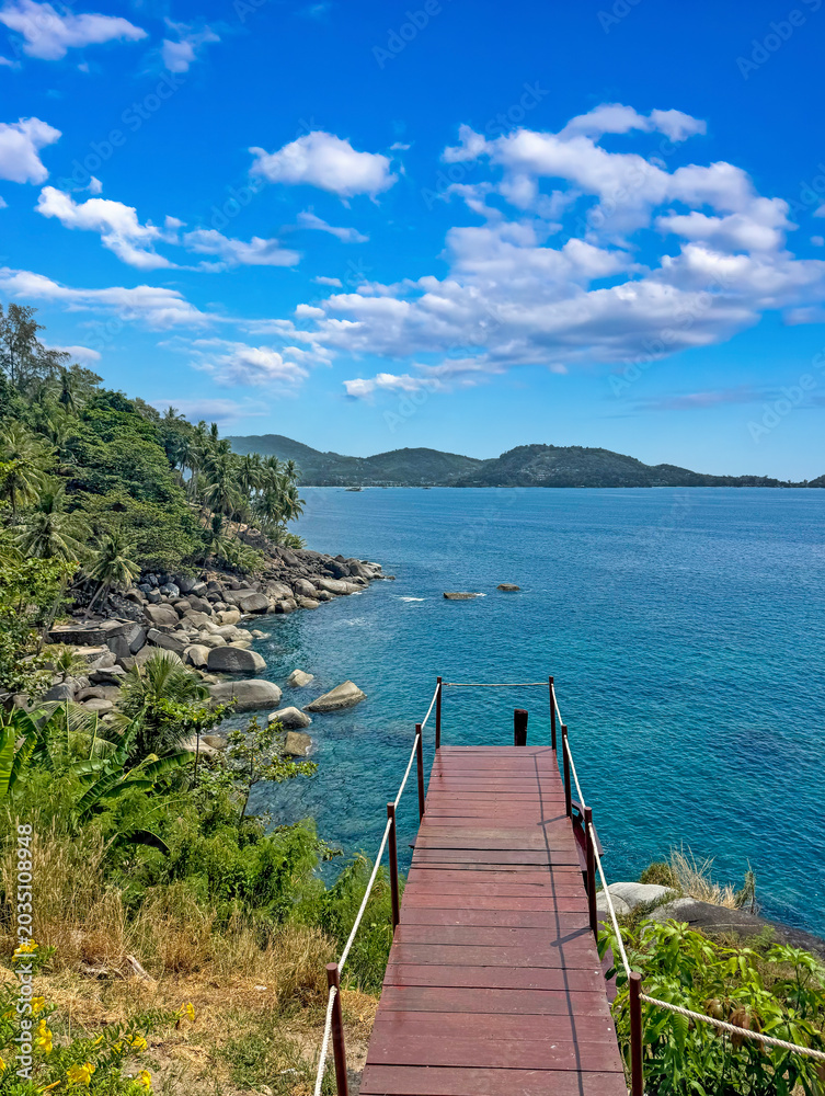 Obraz premium Wooden pier overlooking turquoise tropical sea with rocky coastline palm trees and bright blue sky in Phuket Thailand