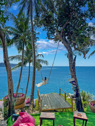 Fotografie Young woman swinging above turquoise tropical sea surrounded by palm trees and r