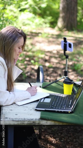 A beautiful 40-year-old female writer works in front of a laptop monitor in a city park. Remote working. Freedom from the confines of a stuffy office in a noisy city. Slow motion vertical 4k footage.