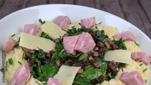 A bowl contains a serving of mushroom and spinach risotto. On top are pieces of Morteau sausage and parmesan cheese. The dish is placed on a wooden surface.