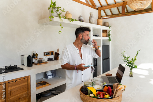Man drinking coffee while checking phone and preparing breakfast in a modern kitchen