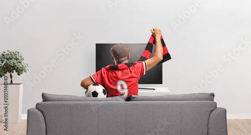 Rear view shot of a senior sports fan in a red jersey cheering with a scarf on a sofa