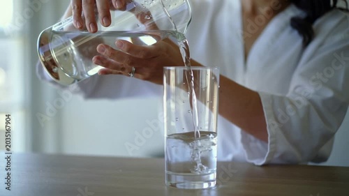 Bringing pitcher, woman tilting, pouring water into tall glass to fill at table, white robe, ring