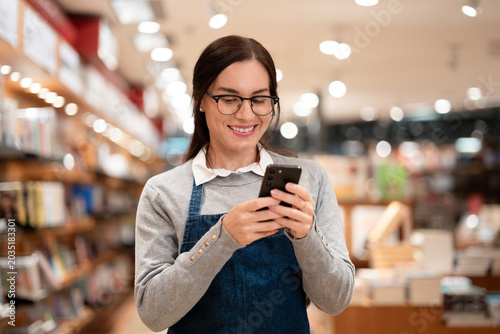 Smiling Latin Employee with Glasses in Gray Sweater and Denim Apron Using Smartphone in Bookstore Retail Store Medium Shot