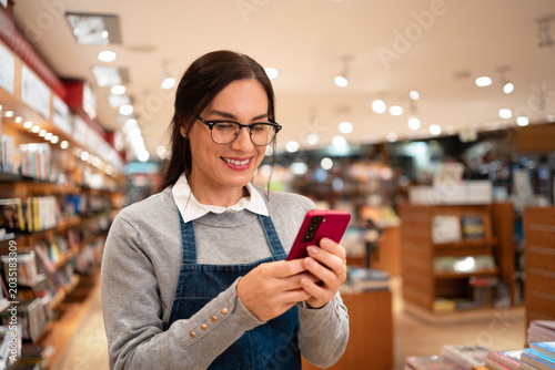Happy Latin Female Worker with Glasses in Gray Sweater and Apron Using Smartphone in Bookstore Retail Store Medium Shot