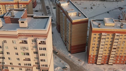 High angle view of modern apartment blocks in winter. Rooftops and ground are covered in fresh snow. Warm sunset light illuminates building facades creating a peaceful, cold scene