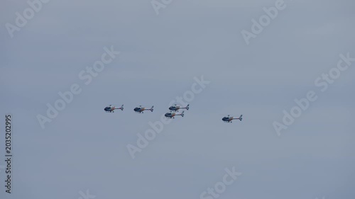 Four light military aerobatic helicopters flying in a loose line formation across a pale grey overcast sky during an air show display