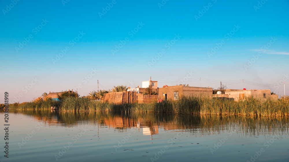 Obraz premium Traditional houses reflect in tranquil waters of Iraqs Marshes at sunset