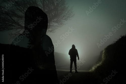 A double exposure of a hooded figure, silhouetted on a spooky foggy road at night