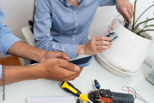 Young Asian woman and man using microcontroller and soil moisture sensors to measure soil moisture in a potted plant.