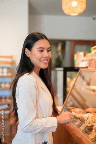 Smiling Thai businesswoman choosing pastries at bakery display counter in cafe.