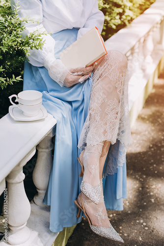 A woman in an elegant blue silk dress, white blouse, and lace gloves sits on a classic stone balustrade, holding a book next to a white porcelain teacup