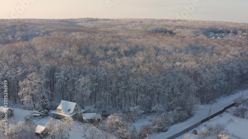 Aerial drone footage flying over a small village with houses and moving towards a vast, dense forest. Trees and ground are covered in fresh white snow during a calm, beautiful winter sunrise