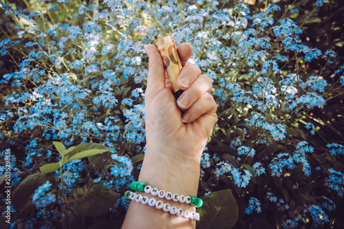 A person wearing a beaded bracelet made of letter beads spelling: money manifestation and holding rolled up 50 euro cash. Blue flower Myosotis alpestris blossoms in the background.