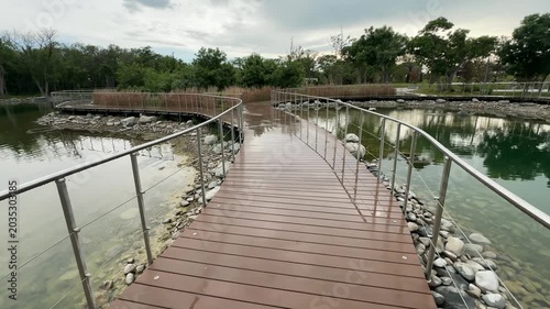 Park boardwalk forward motion over lake, curved wooden path, wet deck, railings, green trees, urban nature, travel recreation.