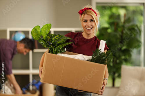 Woman Carries Box of Plants While Man Prepares to Move in a Bright Room During Daytime