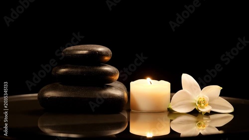 Stack of balanced black zen stones next to a burning candle and a white orchid flower on a reflective dark surface. Concept of harmony relaxation and spa wellness