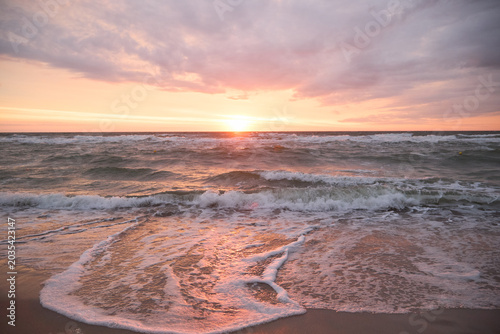 Sea wave with white foam washing onto a sandy beach under a dramatic pink sunset sky.