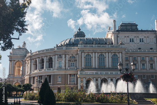 Majestic opera house with ornate facade and domed roof. Lush garden and fountain frame the scene