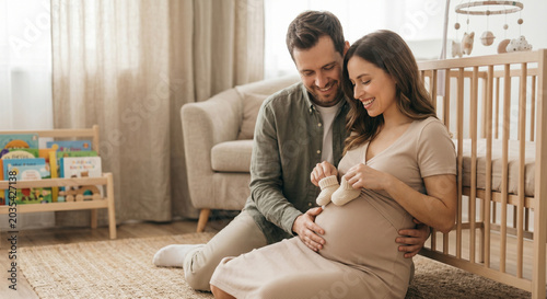 Joyful expectant couple sitting on the nursery floor holding tiny knitted baby shoes against the pregnancy belly.