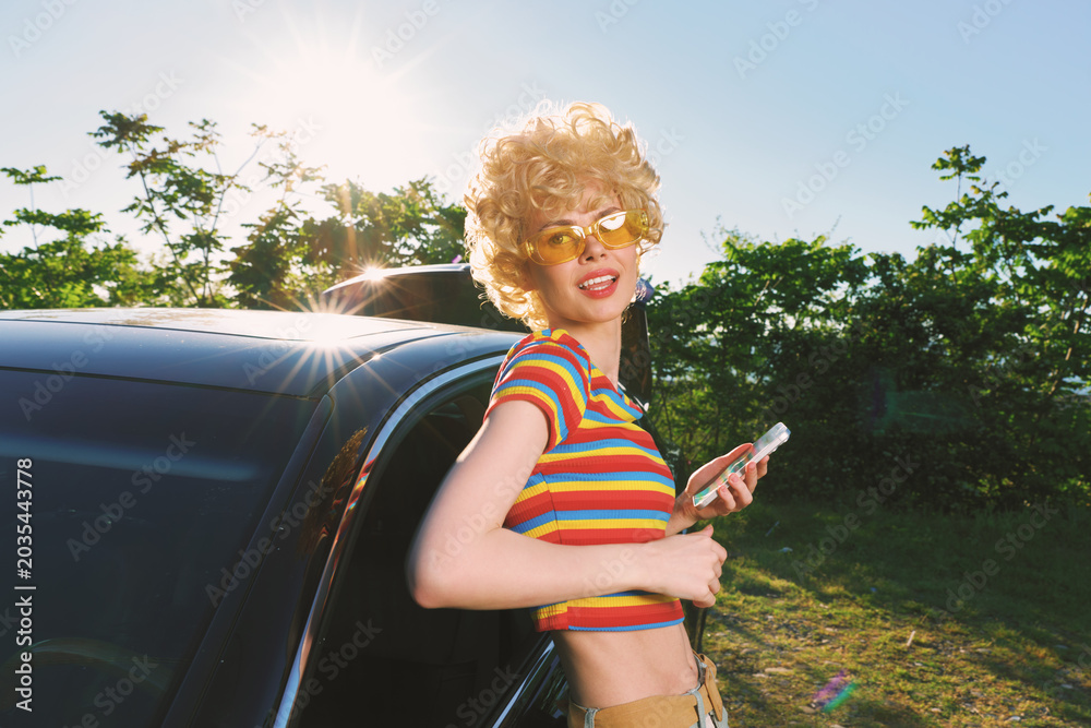 Obraz premium A woman with a retro hairstyle in a striped top stands by a car in bright sunlight, showcasing casual outdoor fashion and a vintage lifestyle moment.