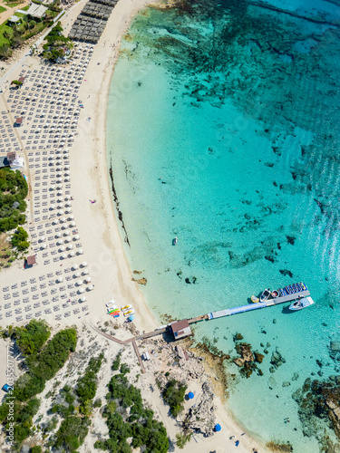 Aerial shot of a Makronissos beach in Ayia Napa, Cyprus, with crystal turquoise water, a wooden pier,