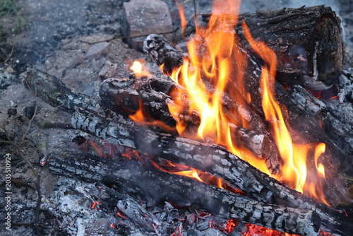 Bright bonfire with orange flames engulfing charred wooden logs. Atmospheric view of evening campfire with smoke and sparks outdoors. Burning wood in bonfire, hot coals