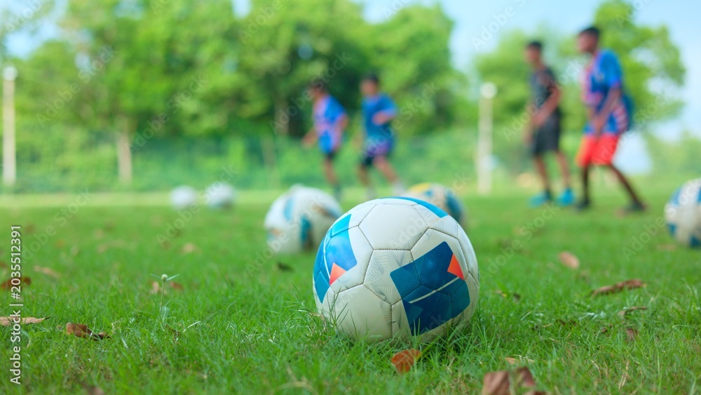 Fototapeta premium Close up of a soccer ball on green grass with blurred children training in the background during summer sports camp.
